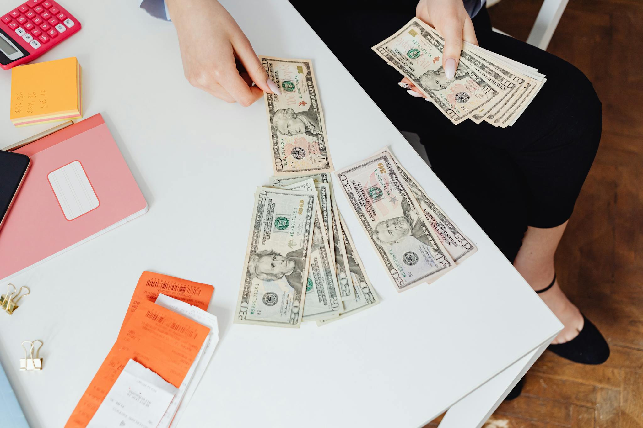 A person counting US dollar bills at a white desk with office supplies nearby.